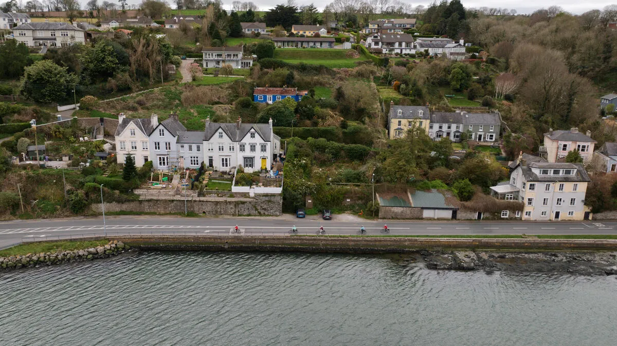 Aerial view of cyclists riding on a road next to water, with houses and gardens on the hillside.