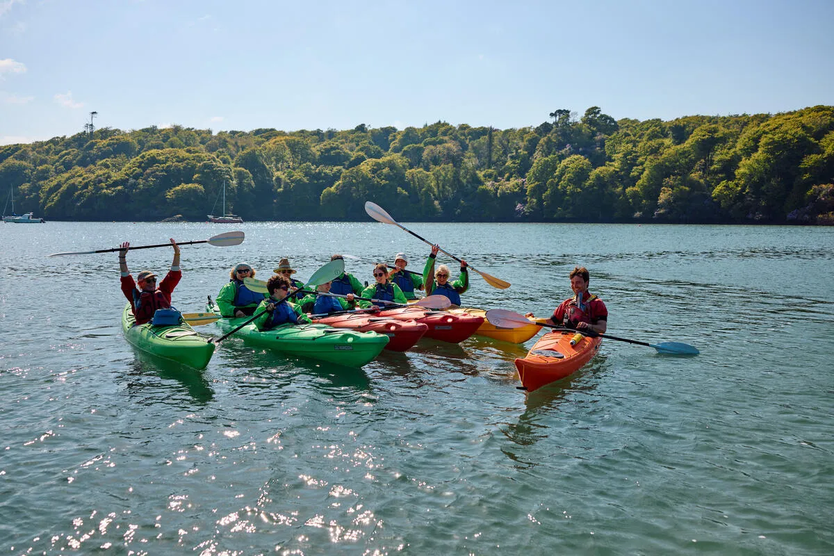 A group of people in colorful kayaks paddling on the water with a forested shoreline in the background.