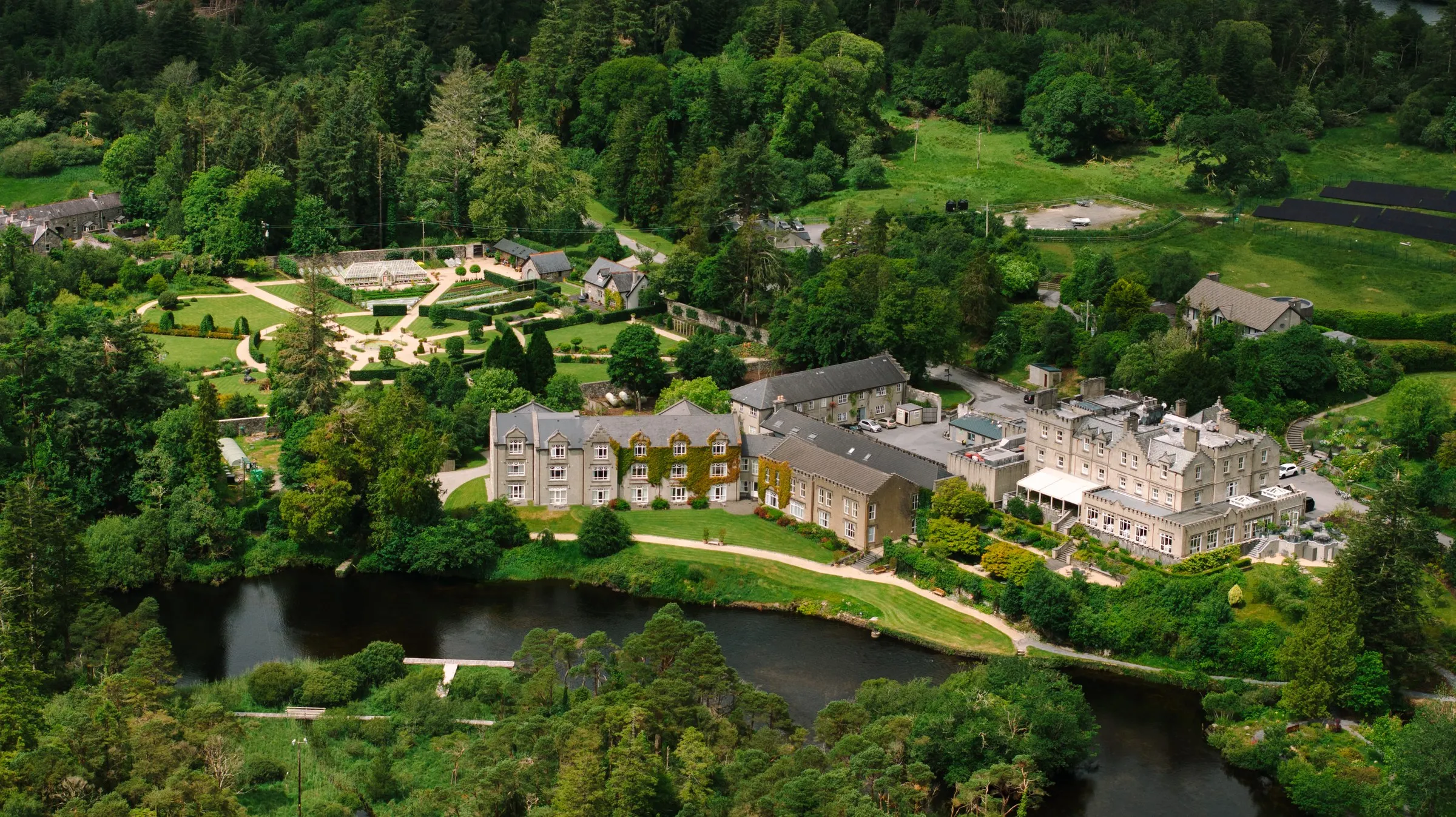 Aerial shot of Ballynahinch Castle Hotel, a large stone building, by a dark river, surrounded by dense green forests.