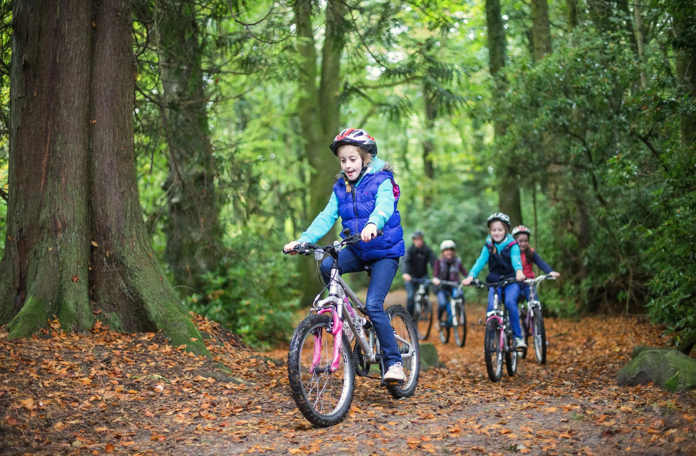 A girl in a blue vest leads children riding bikes on a leaf-covered forest trail.