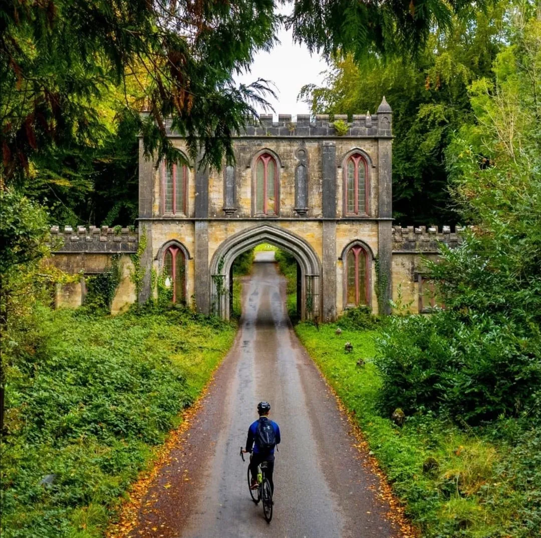 A cyclist on a road approaching a large, ancient stone archway surrounded by lush green trees and foliage.