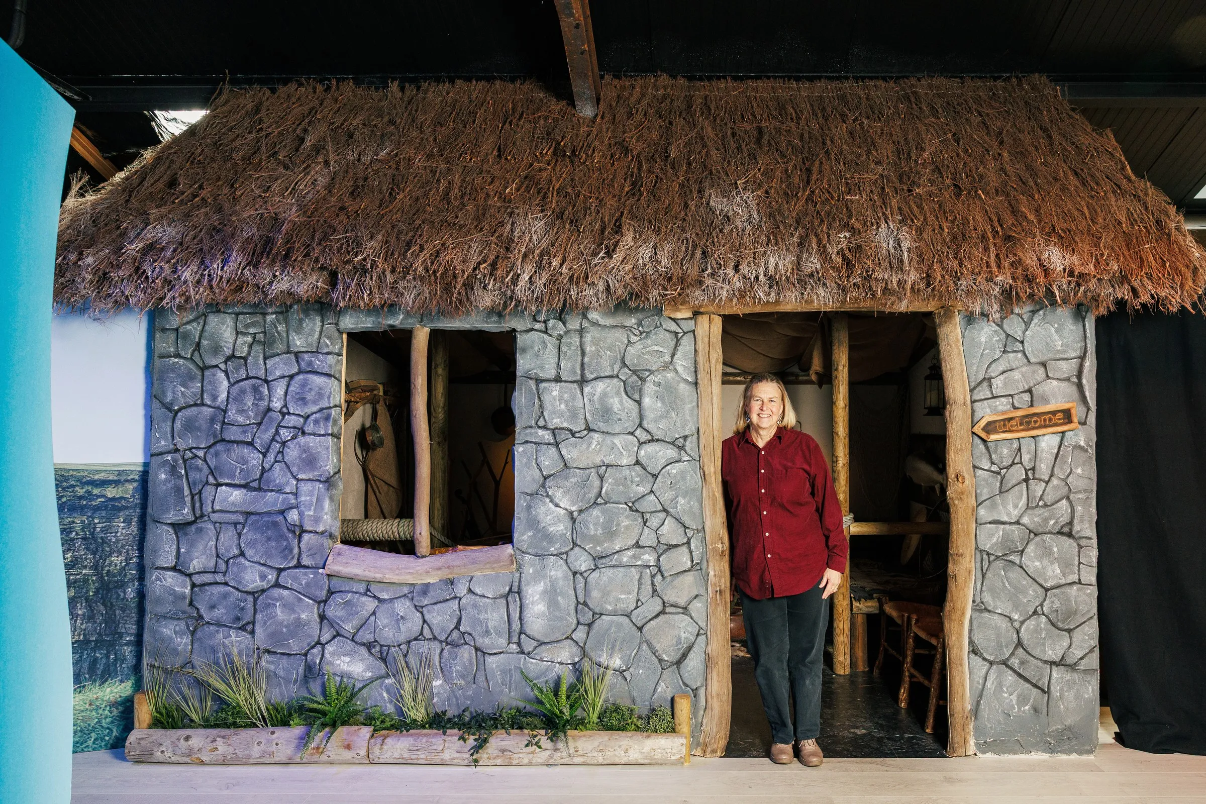 A woman stands in the doorway of a stone cottage with a thatched roof and a welcome sign.