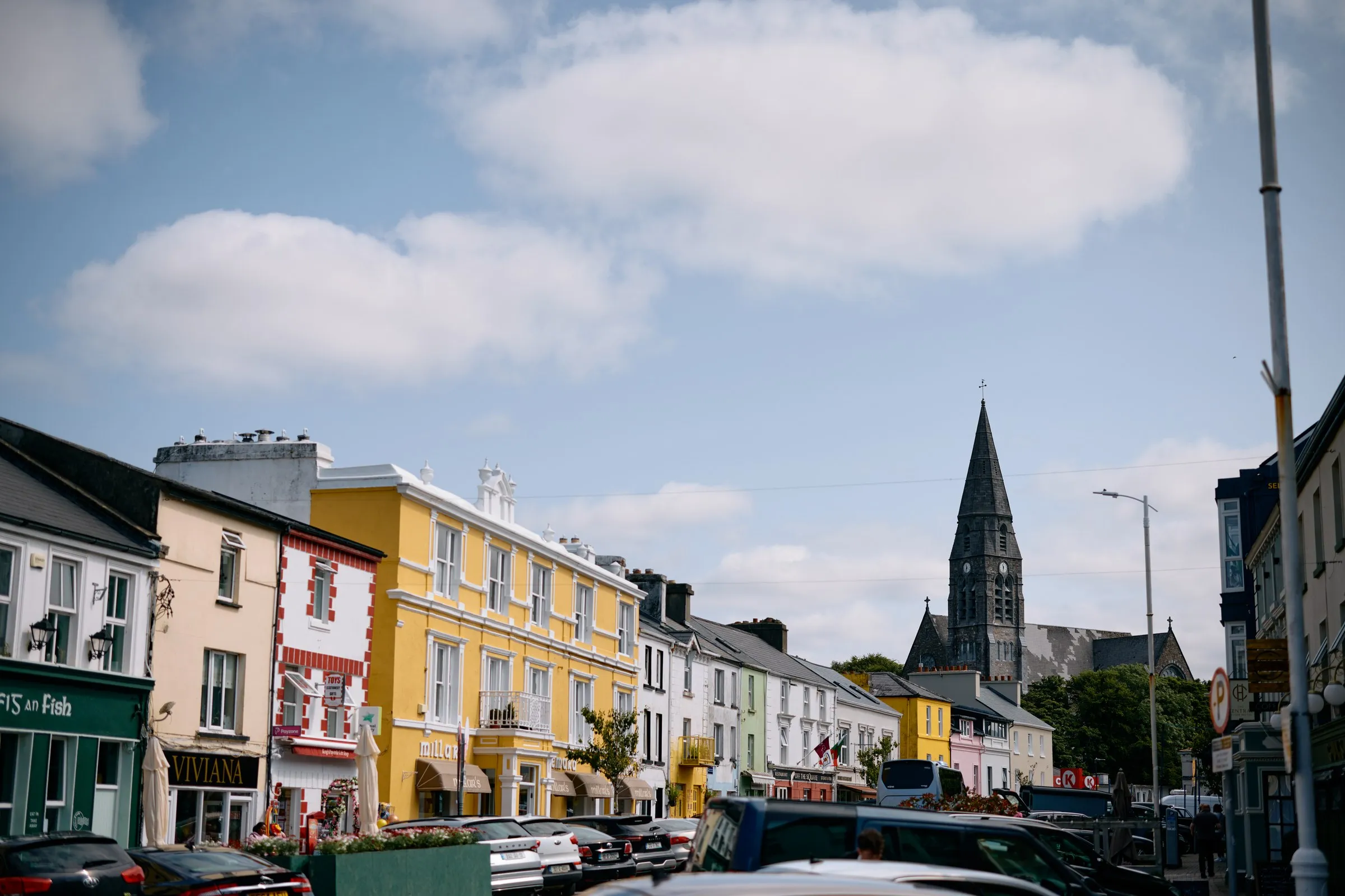 A street scene in Clifden with colorful buildings, parked cars, and a tall church steeple under a blue sky.