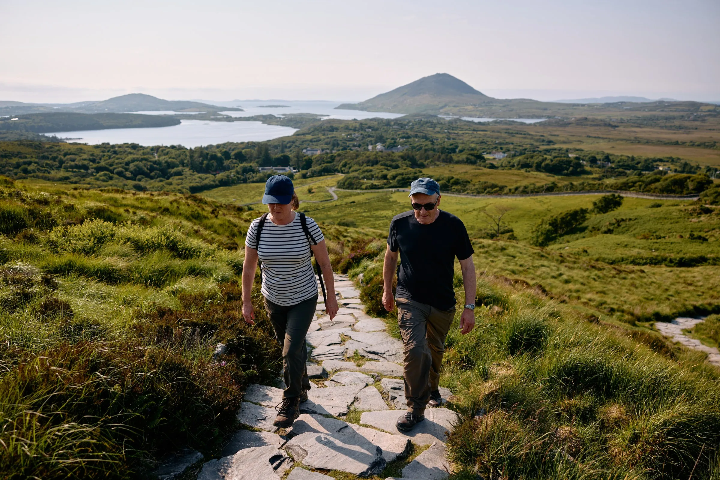 Two hikers ascend a stone path, with green hills, lakes, and distant mountains in Connemara.