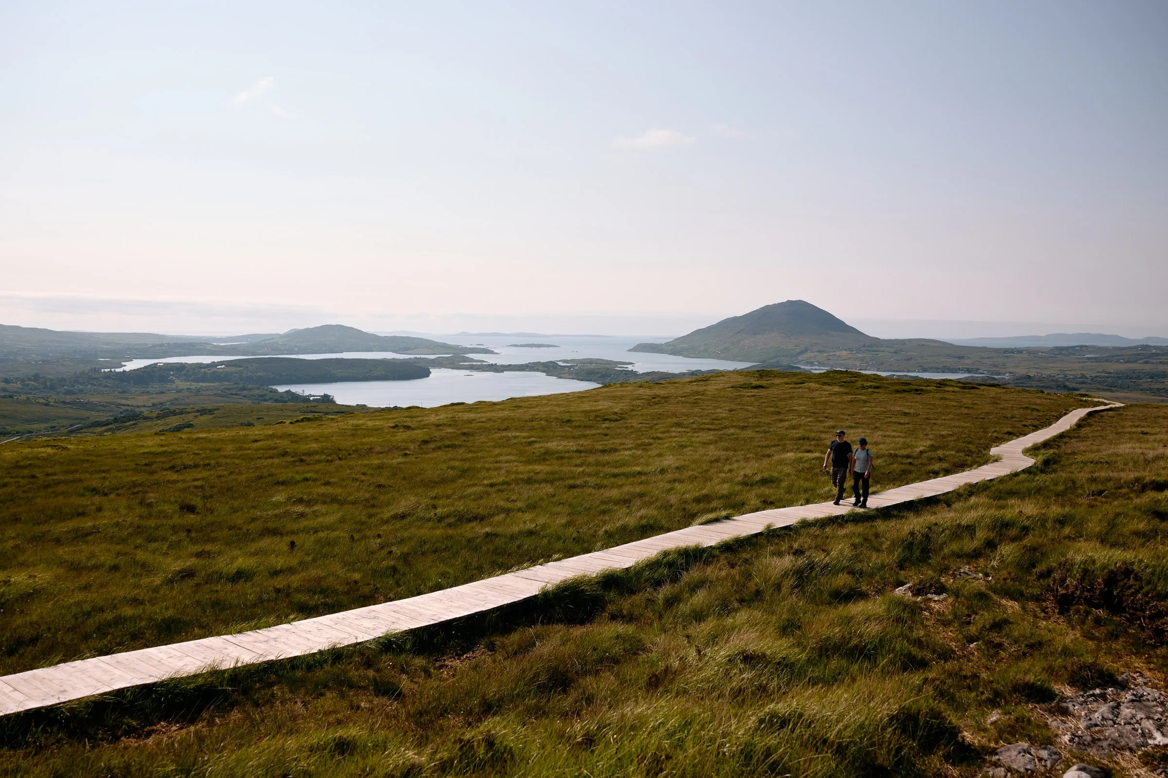 Two people walk on a wooden boardwalk through a grassy landscape with lakes and hills under a clear sky.