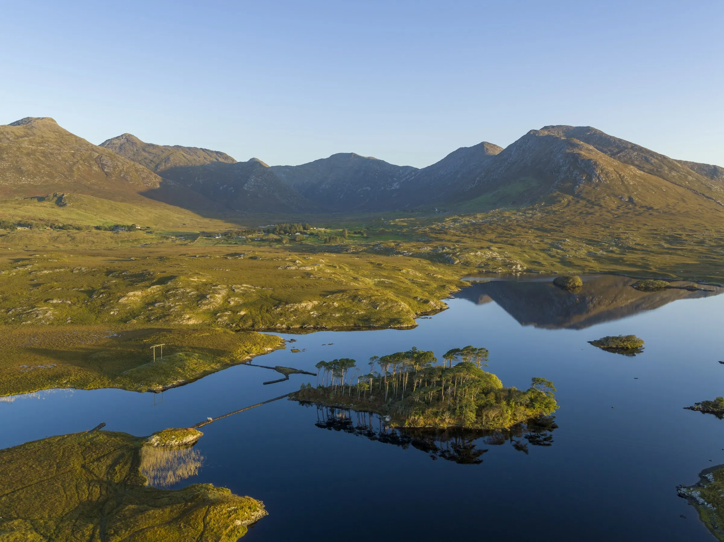 Aerial view of Derryclare Lough with a tree-covered island and causeways, set against the Twelve Bens mountains.