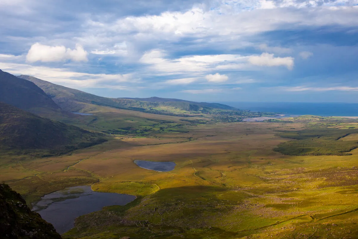 High angle view of a vast valley with small lakes, brown and green terrain, and mountains under a cloudy sky.