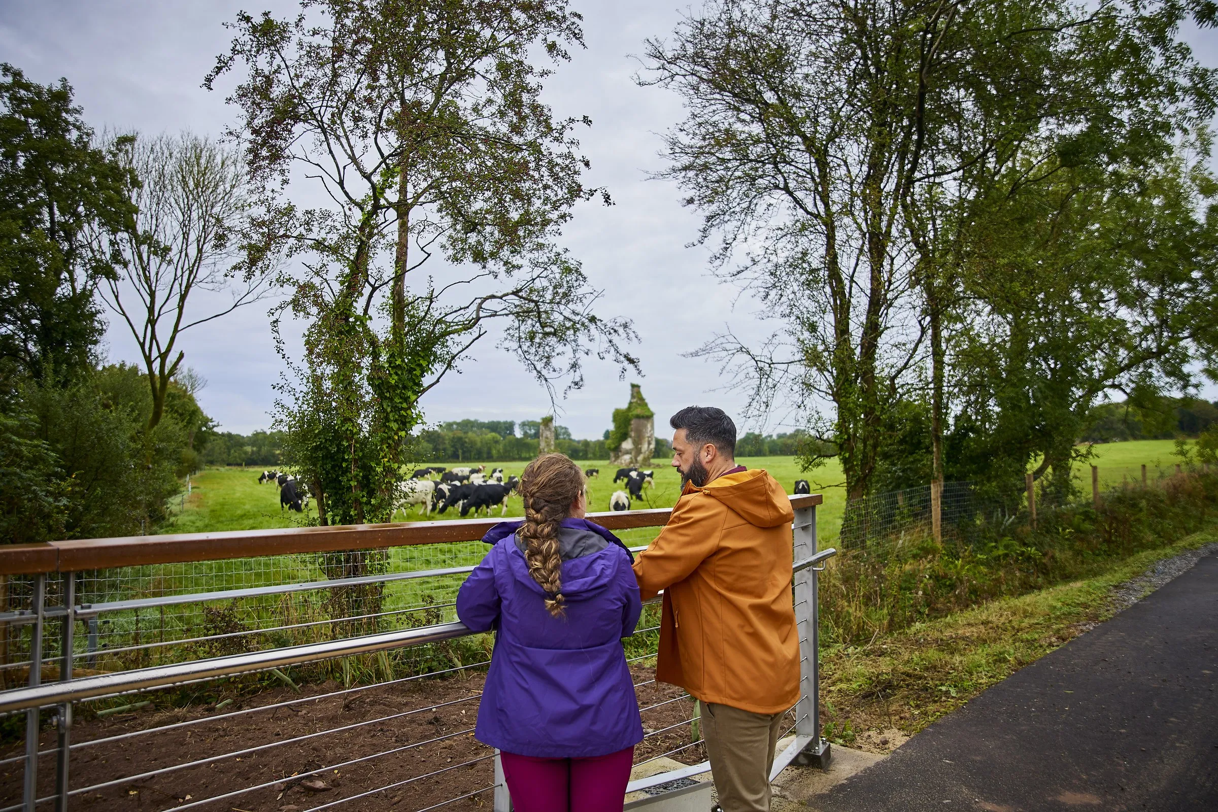 A couple leans on a railing overlooking a pasture with cows and stone ruins in the distance.