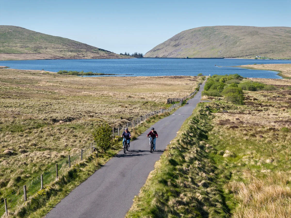 Two cyclists ride down a paved road with grassy fields and a blue reservoir in the background.