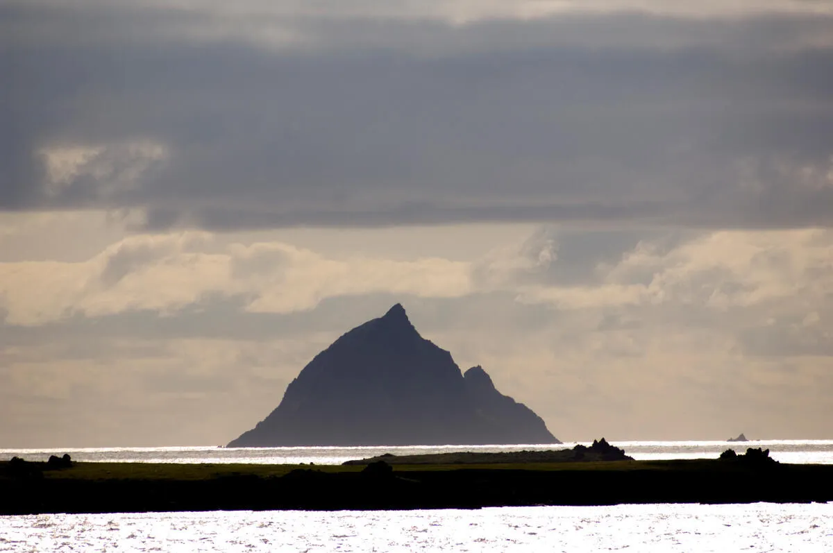 Silhouette of Tearaght Island rising from the sea with a grassy foreground and cloudy sky above.