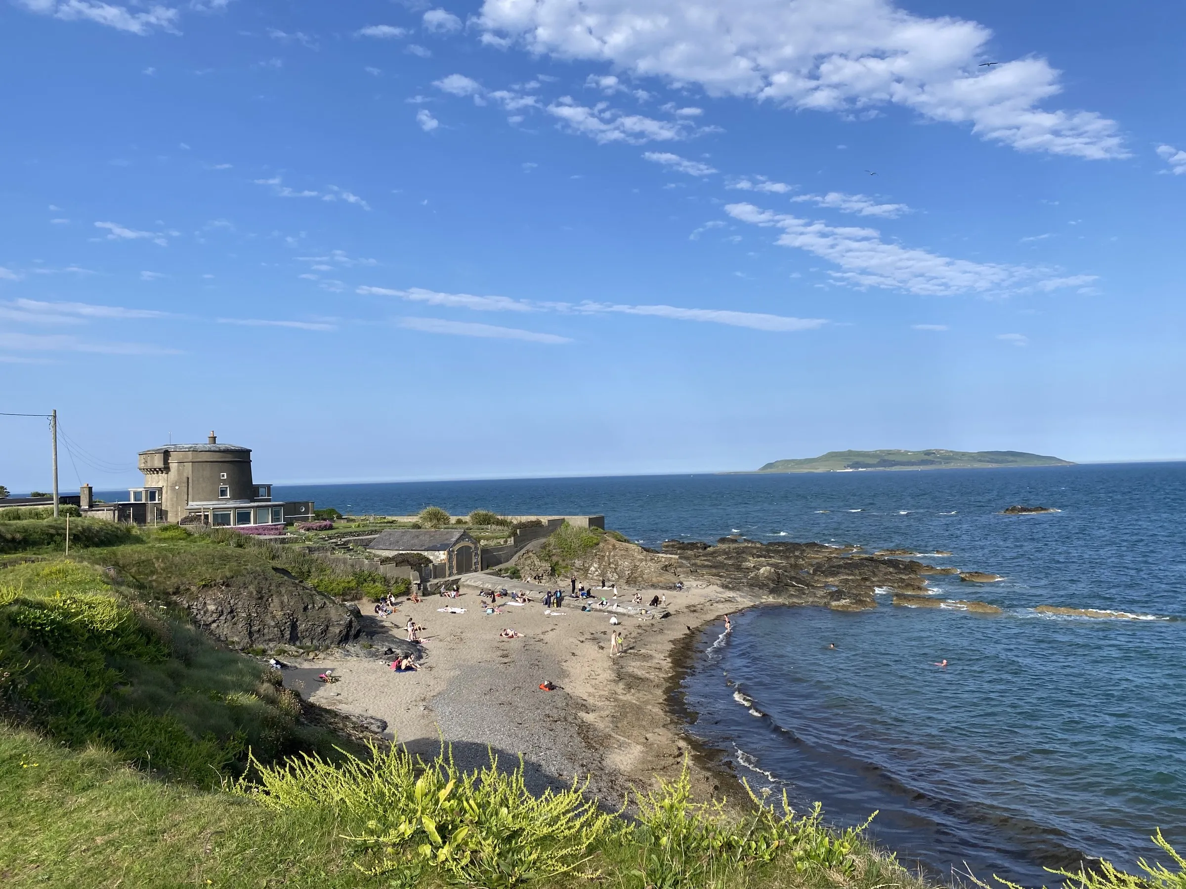 A Martello tower and a sandy beach with people, with an island in the distance under a blue sky.