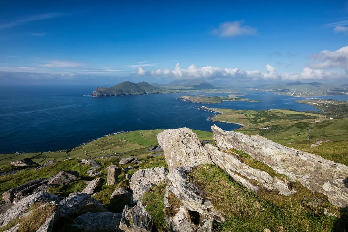 Large rocks in the foreground overlooking a blue bay, an island, and green coastal hills.