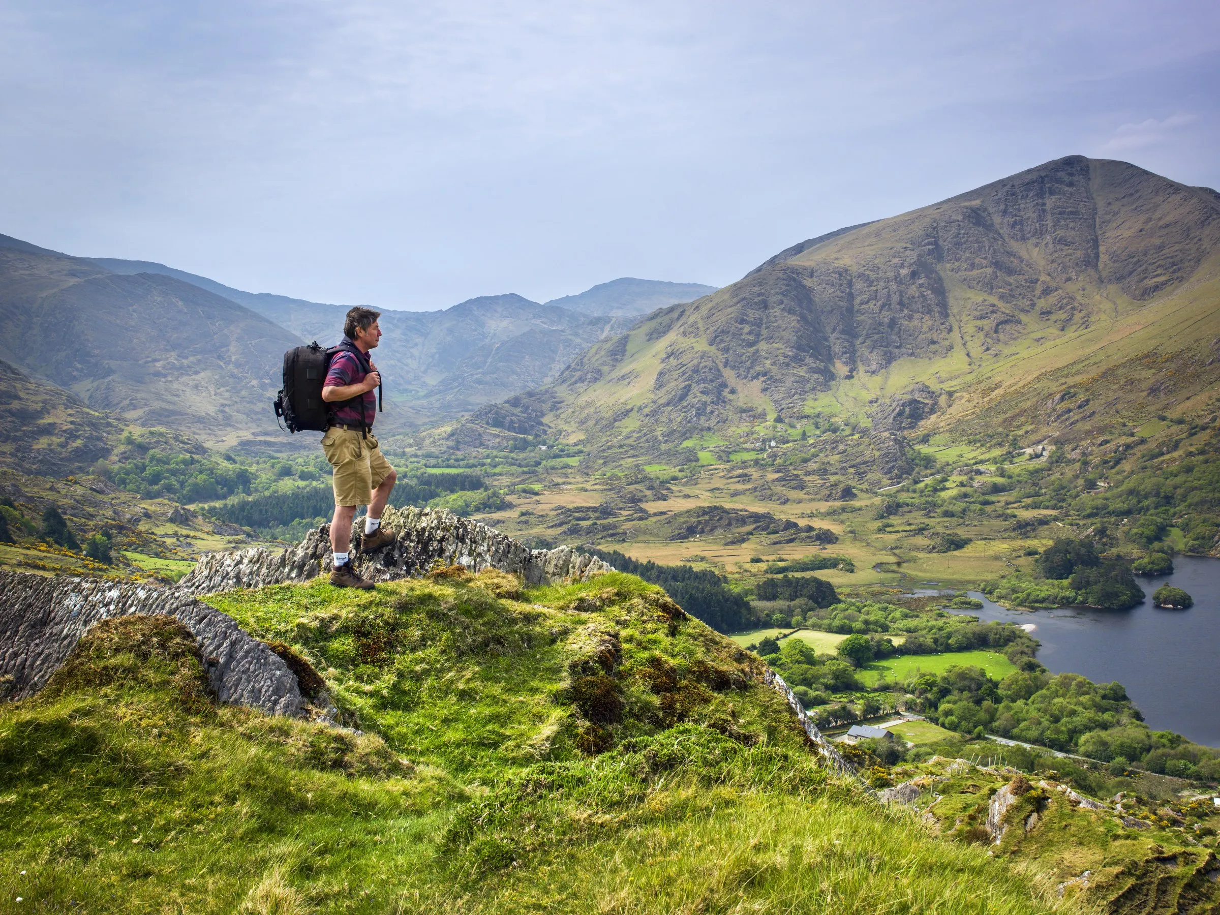 A hiker with a backpack stands on a rocky ridge overlooking a green valley and a lake.