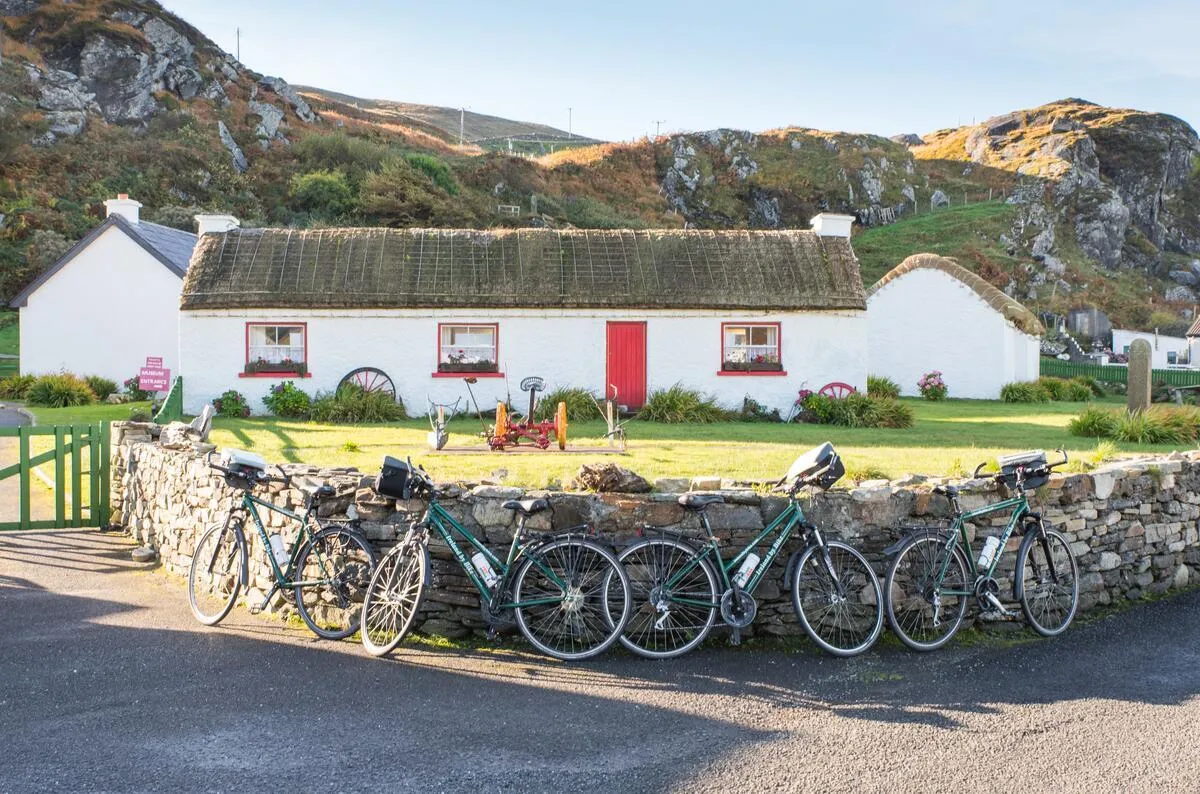 Four green bicycles lean against a stone wall in front of a white thatched cottage with a red door.