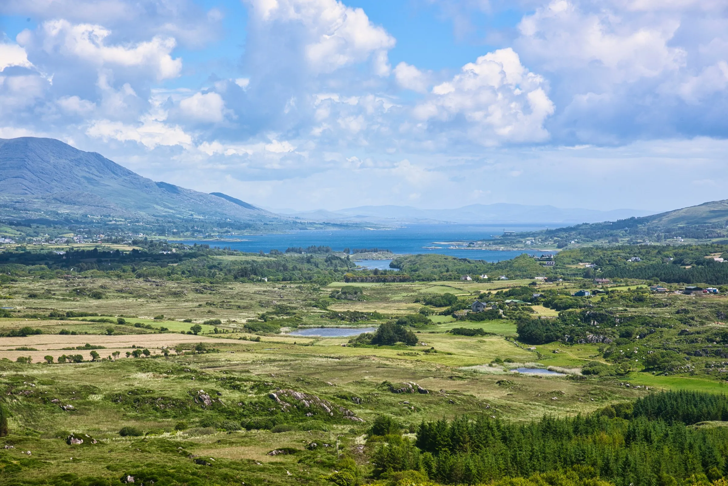 A wide landscape view of green fields, distant mountains, and a blue bay under a cloudy sky.