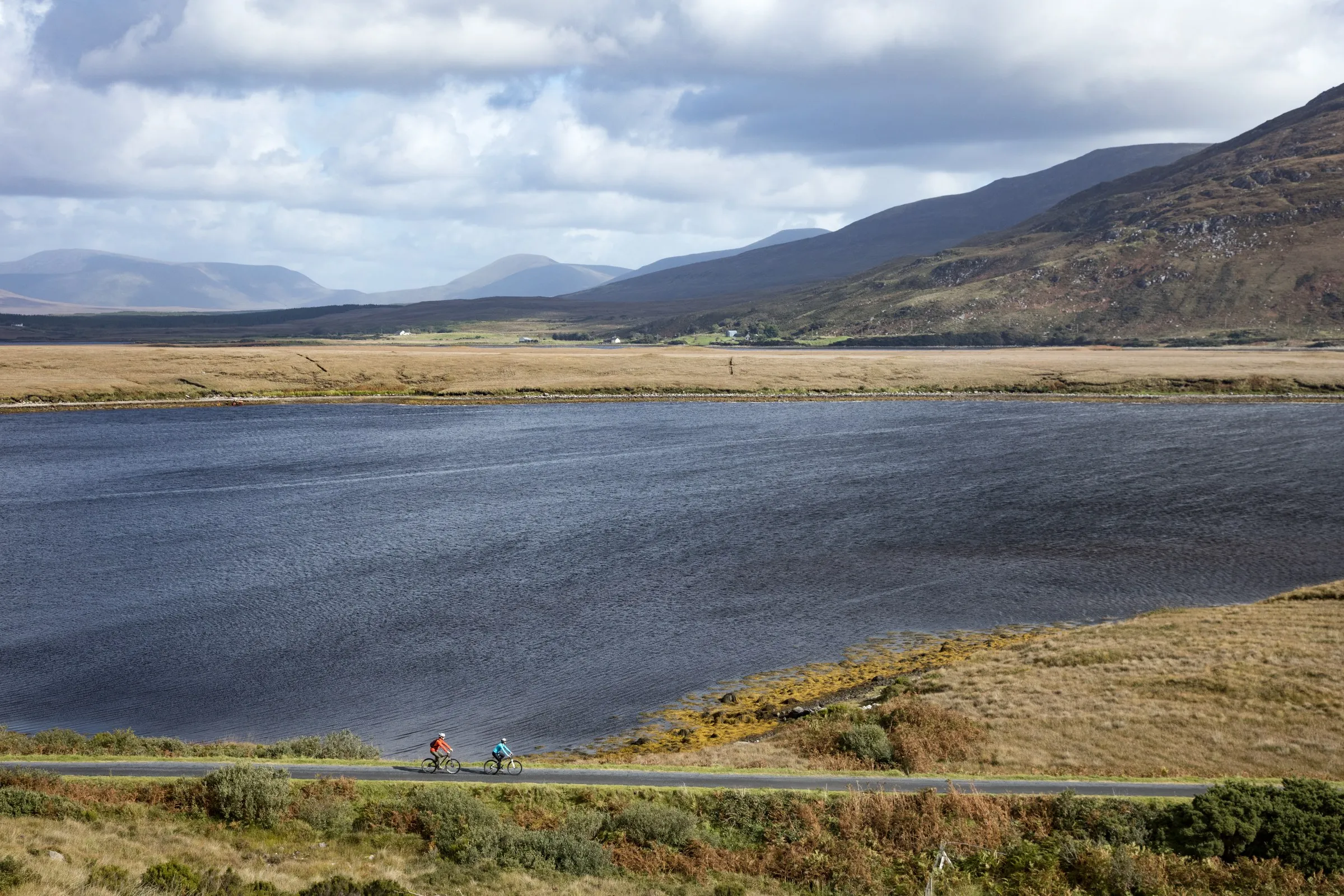 Two cyclists ride on a path in the foreground with a large bay and mountains in the distance.