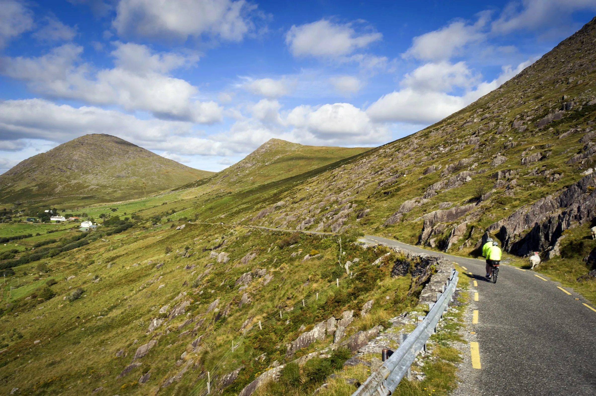 Two cyclists in yellow jackets ride up a winding mountain road with rocky green slopes and a blue sky.
