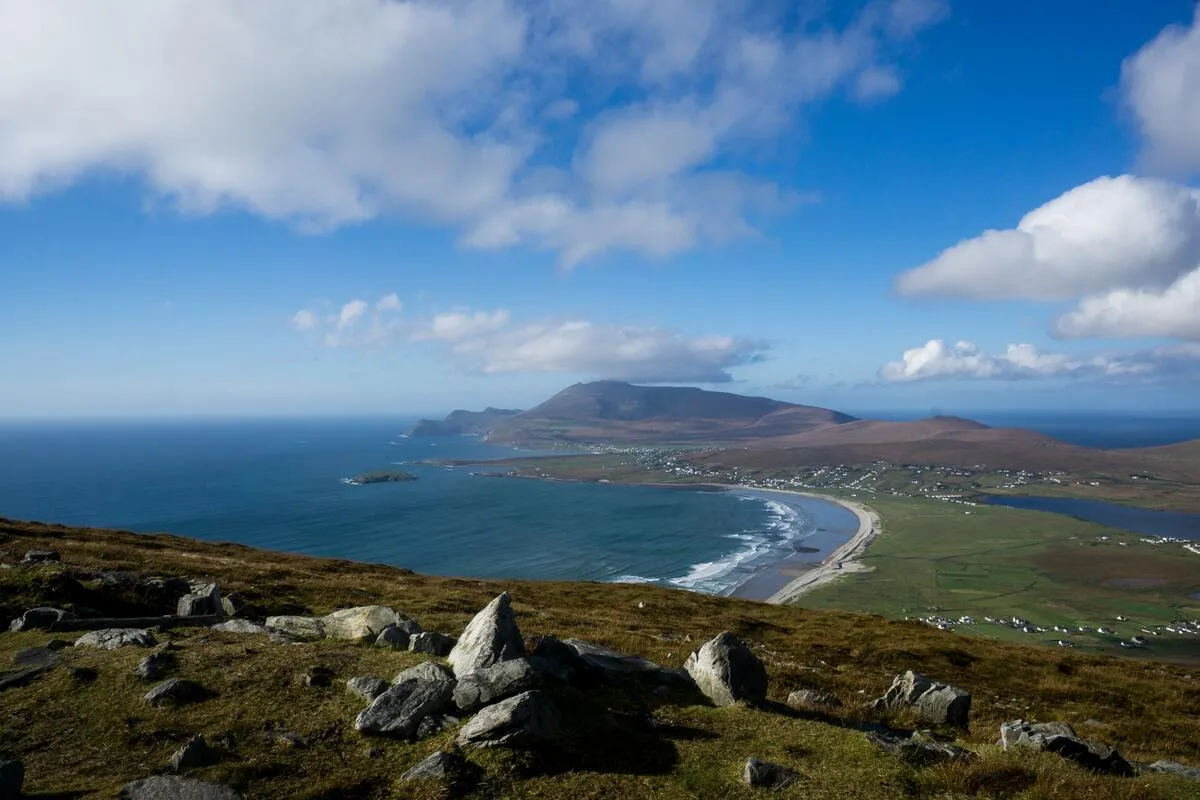 A rocky foreground overlooking Keel Beach, a long sandy shoreline, green fields, and the blue Atlantic Ocean.