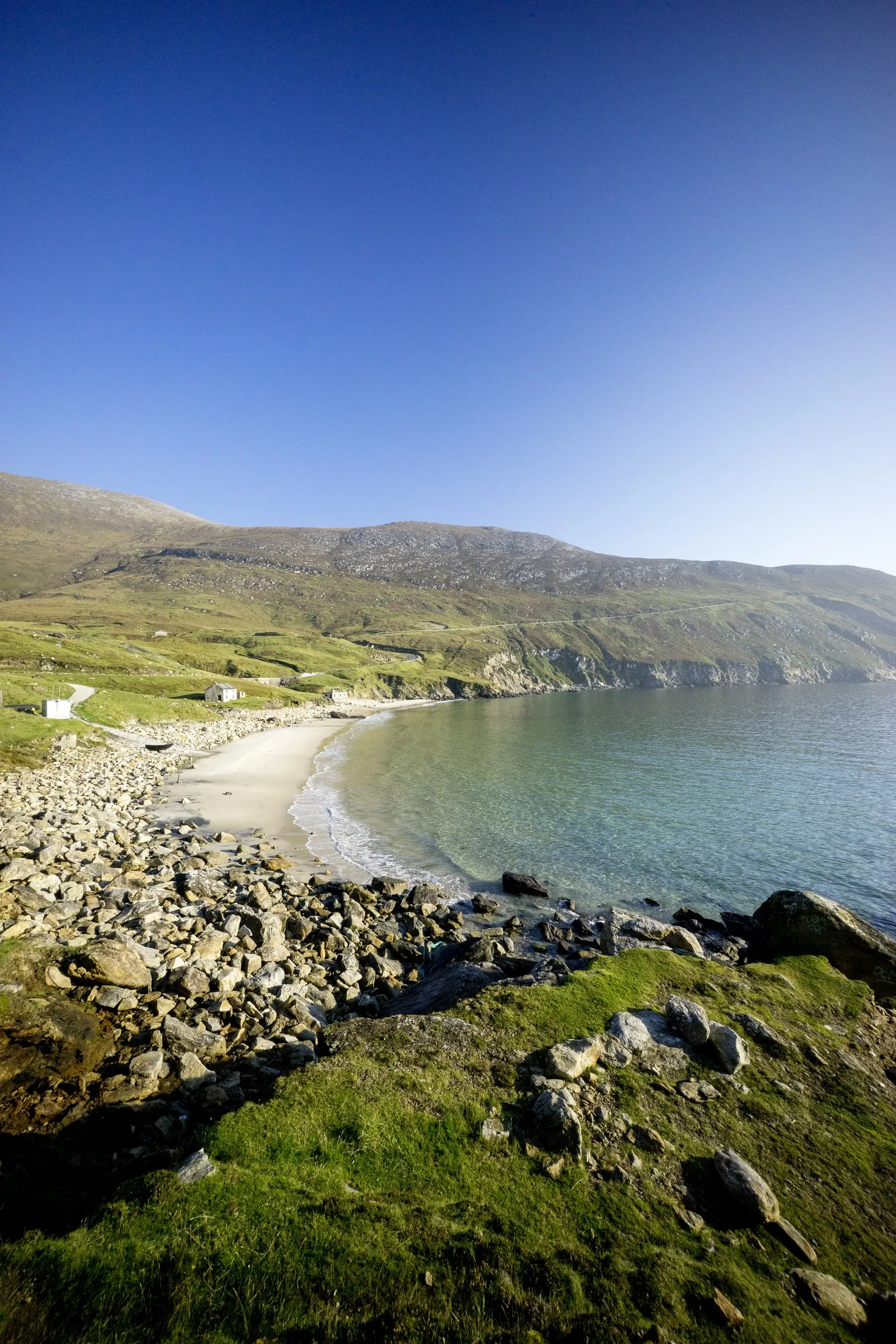 A rocky foreground overlooks a sandy beach curving along a calm bay with green hills in the background.