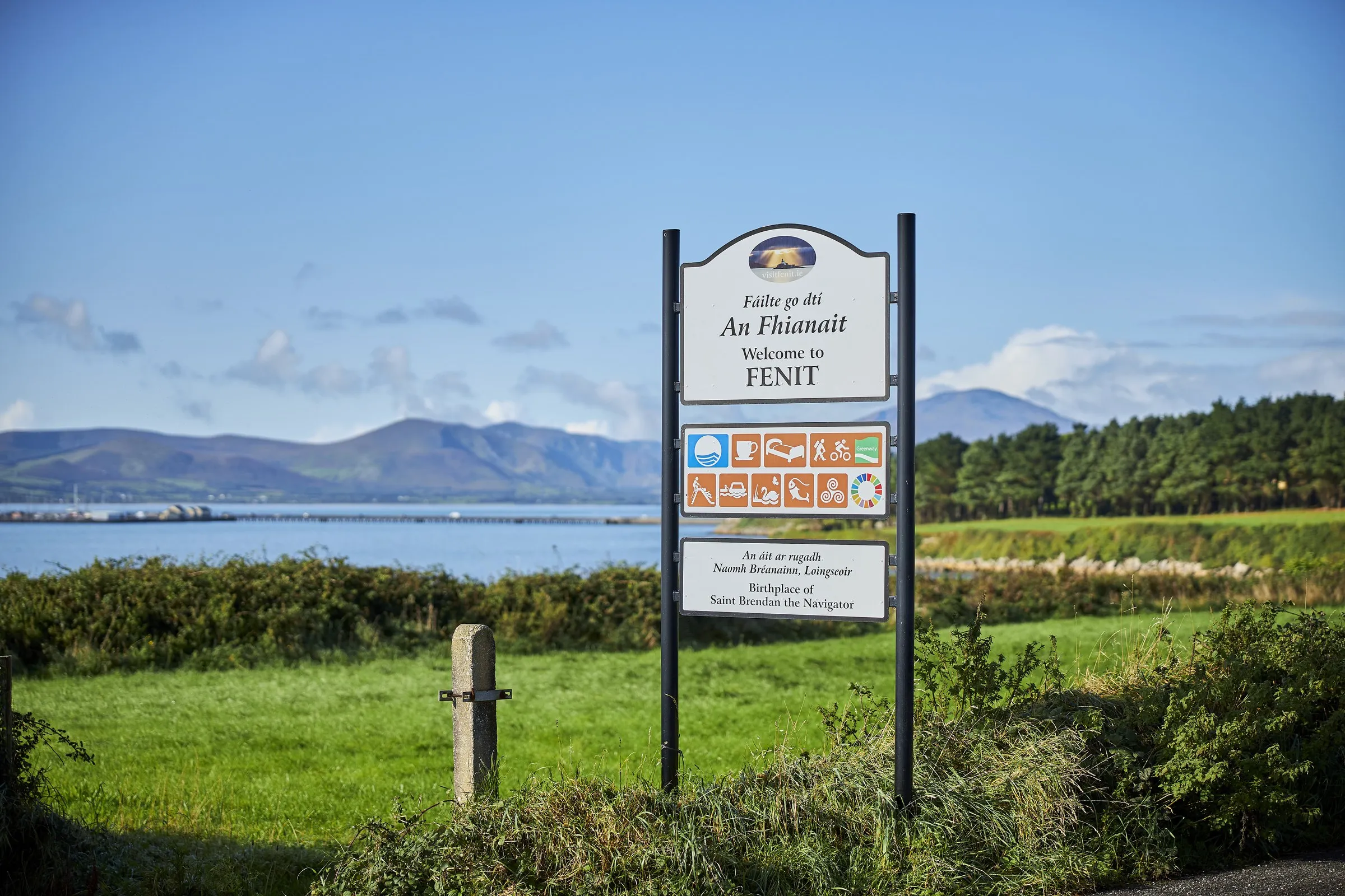 Signpost for Fenit on the Kerry Greenway stands in a grassy field with mountains and water in the background.