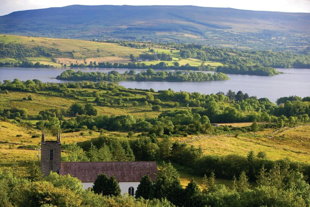 A stone church sits on a grassy hill overlooking a large lake with tree-covered islands and mountains.