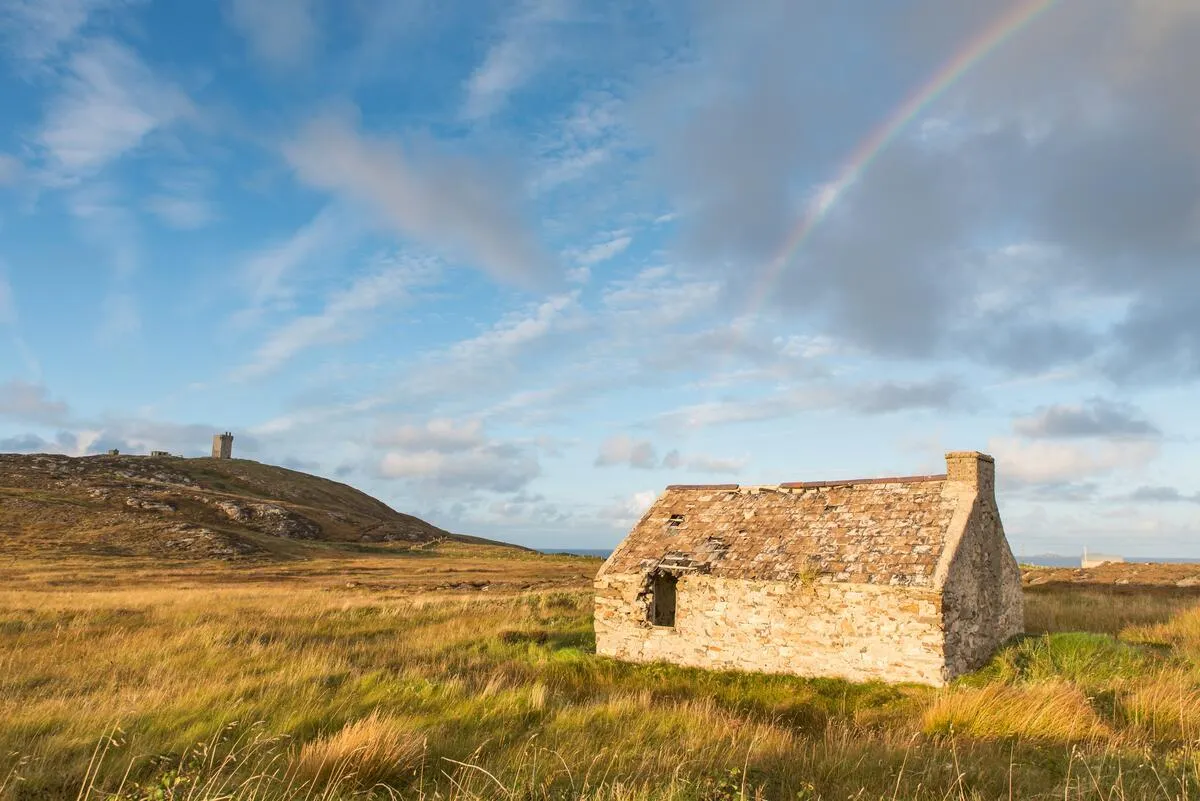 A stone ruin stands in a grassy field with a rainbow arching across a blue sky.
