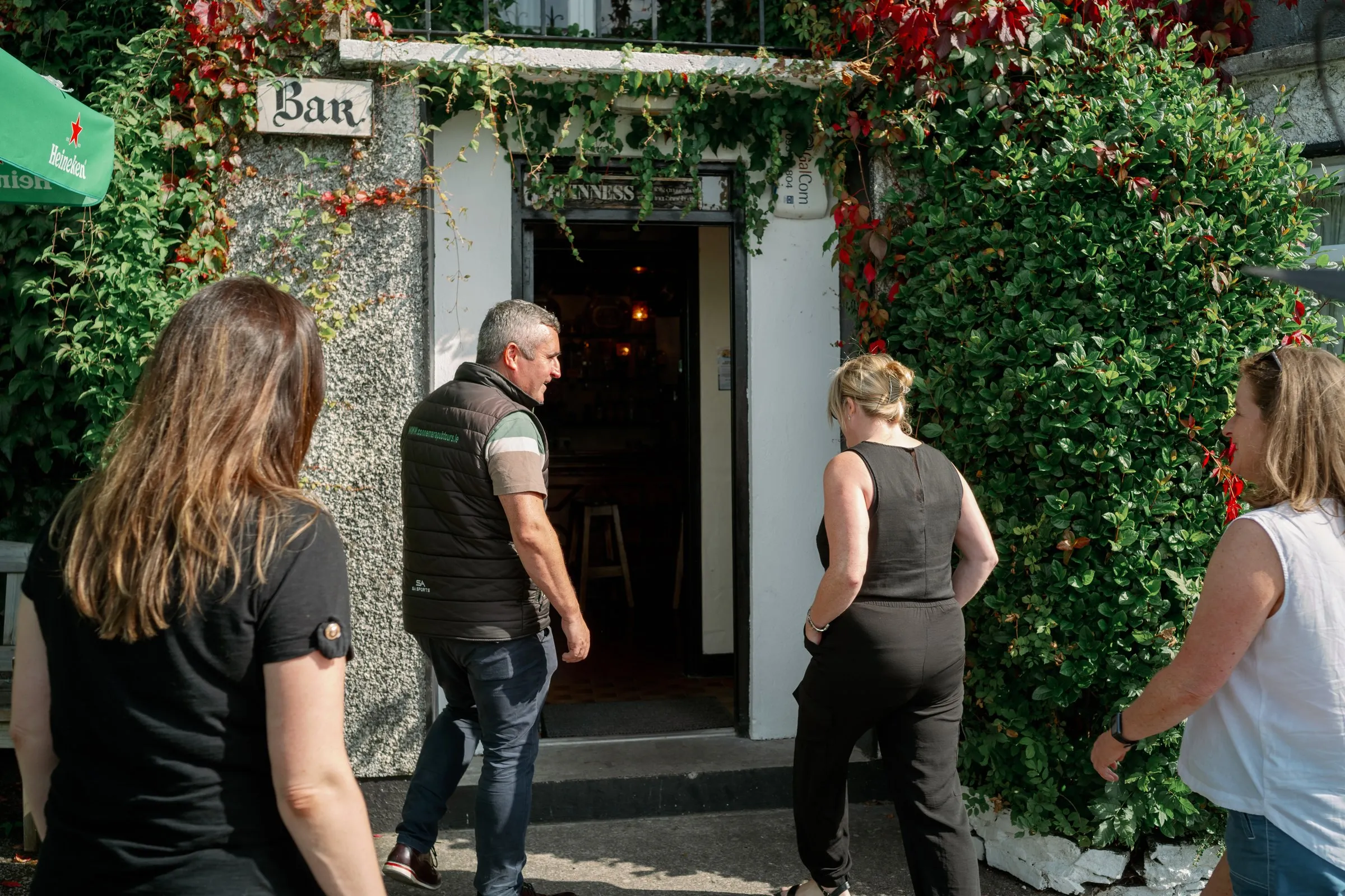 A tour guide talks to three women outside a pub entrance covered in green ivy.