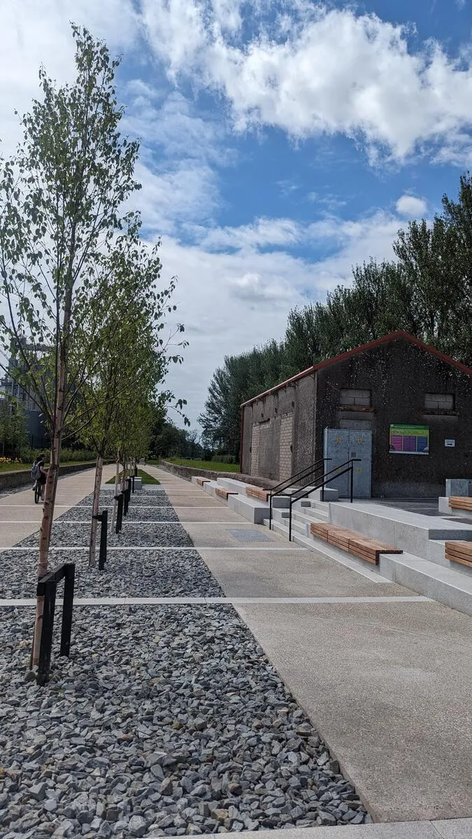 A paved path runs alongside a gravel strip with young trees, next to a building with tiered concrete benches.