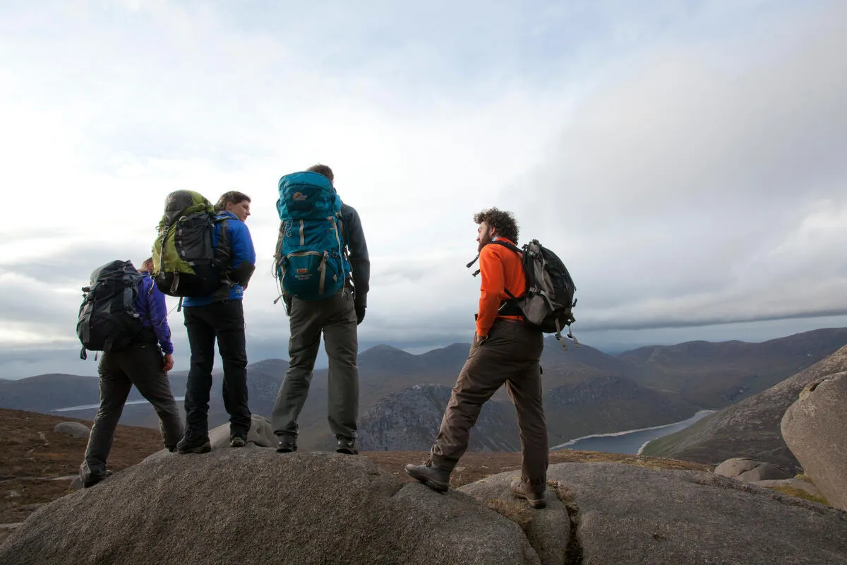 Four hikers with backpacks stand on a large grey rock looking out at distant mountains and a lake.