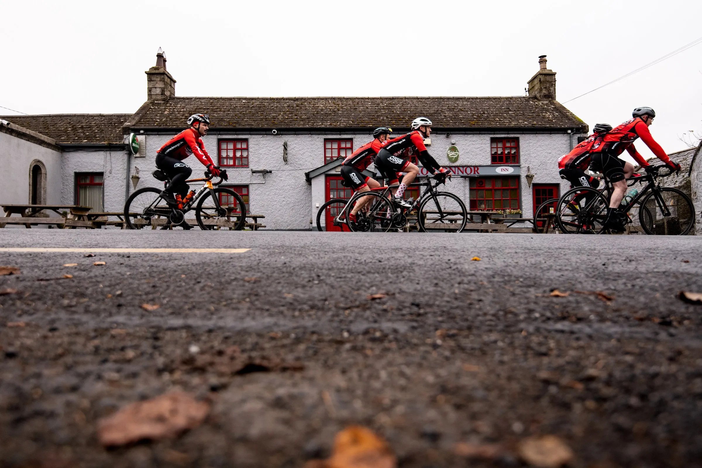 Five cyclists in red and black jerseys ride bicycles past a white stone building with red windows.