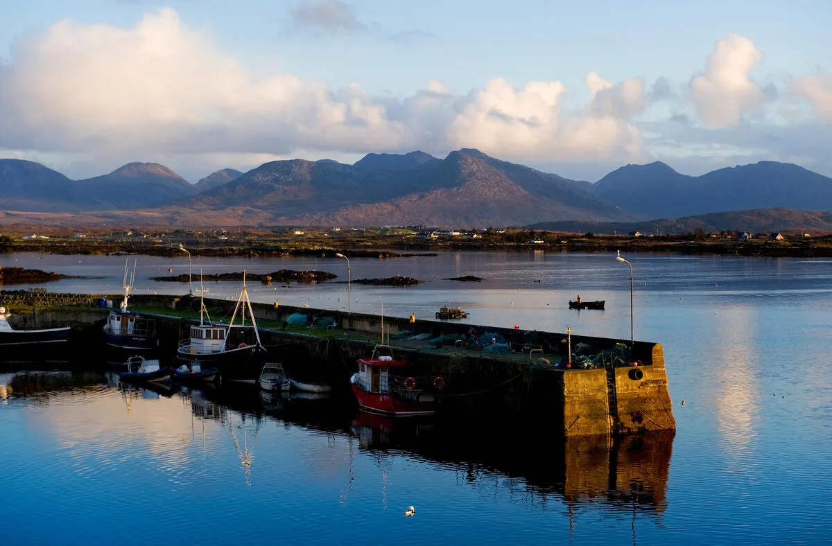 Fishing boats docked along a stone harbor wall in Roundstone with calm water and mountains in the background.