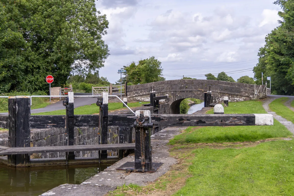 Wooden canal lock gates and a metal winch stand before a stone arch bridge crossing the water.
