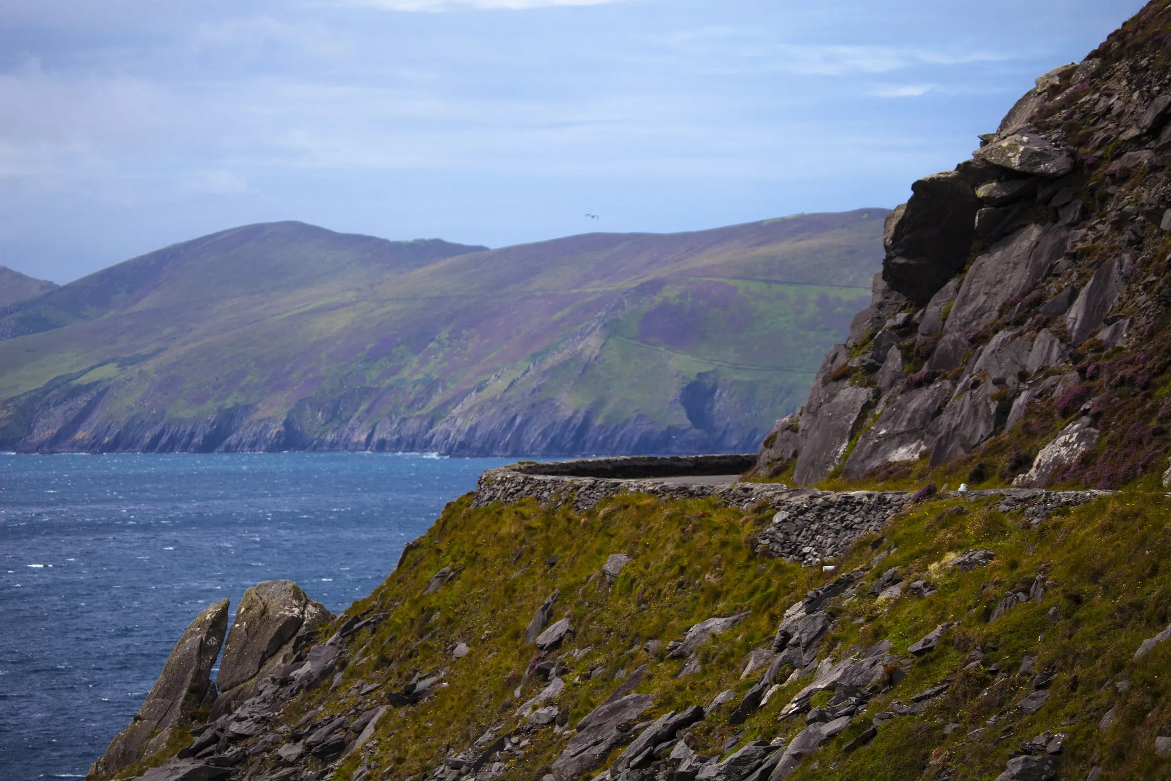 A stone wall borders a grassy cliff edge overlooking the blue Atlantic Ocean and distant mountains.