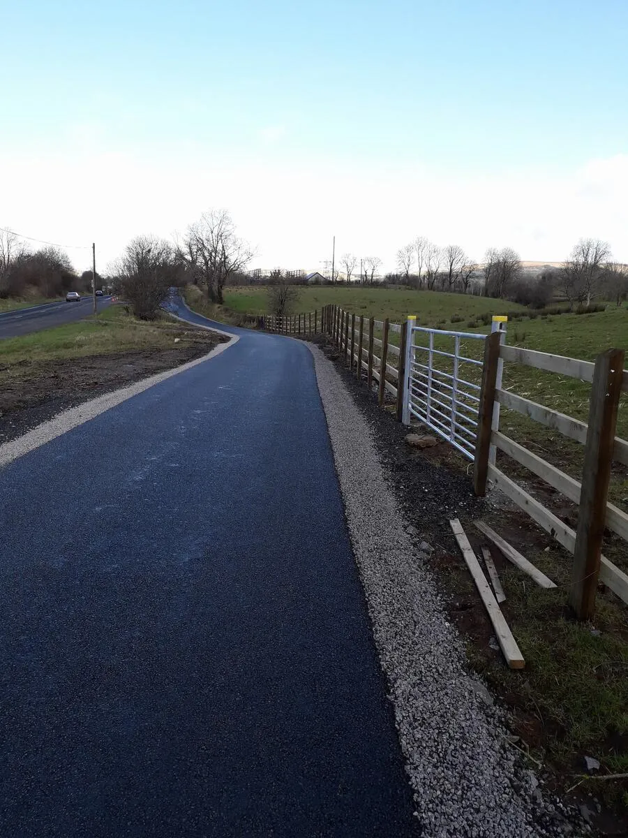 A paved asphalt path curves alongside a wooden fence and metal gate in a rural landscape.