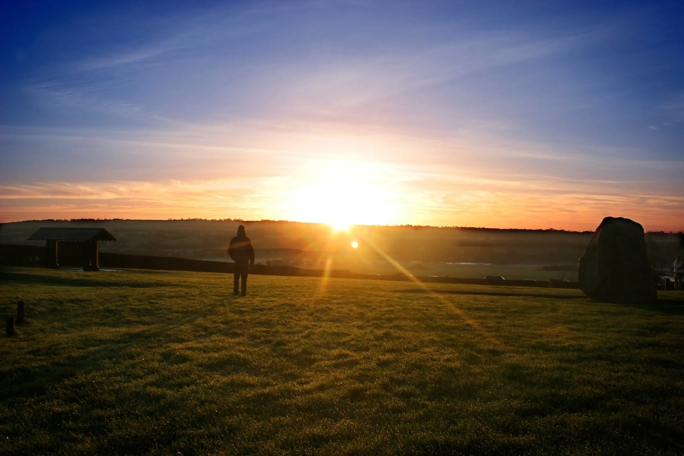 A person stands on a grassy field facing a bright sunrise with a large standing stone on the right.