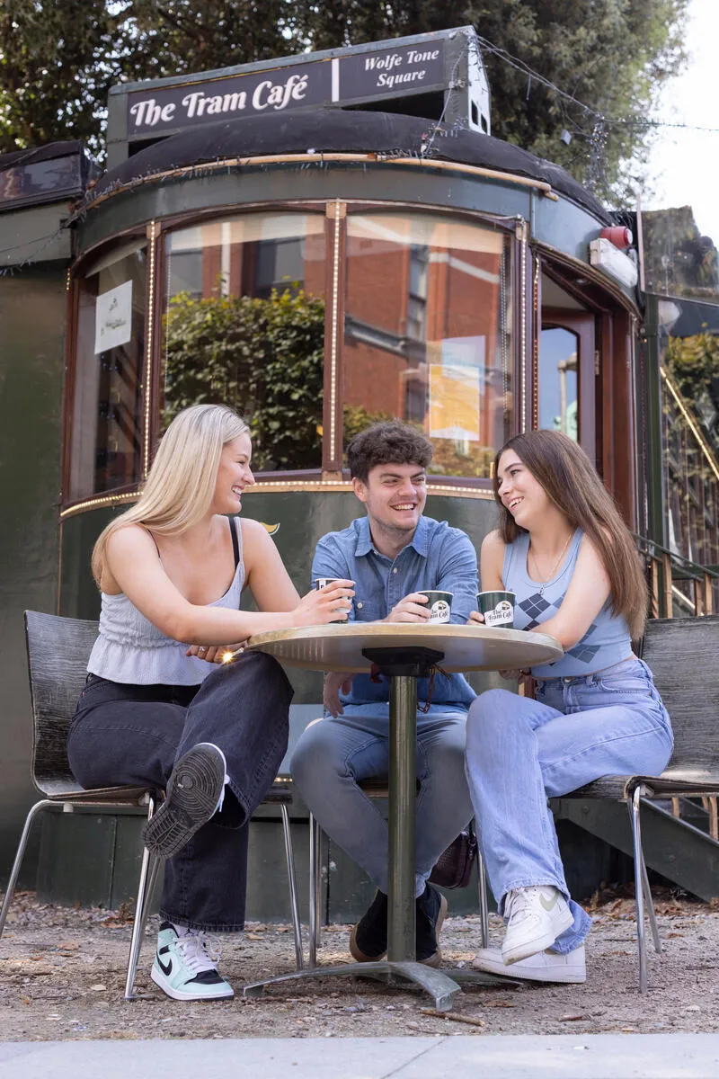 Two women and a man sit outside a vintage tram cafe, holding coffee cups and talking.