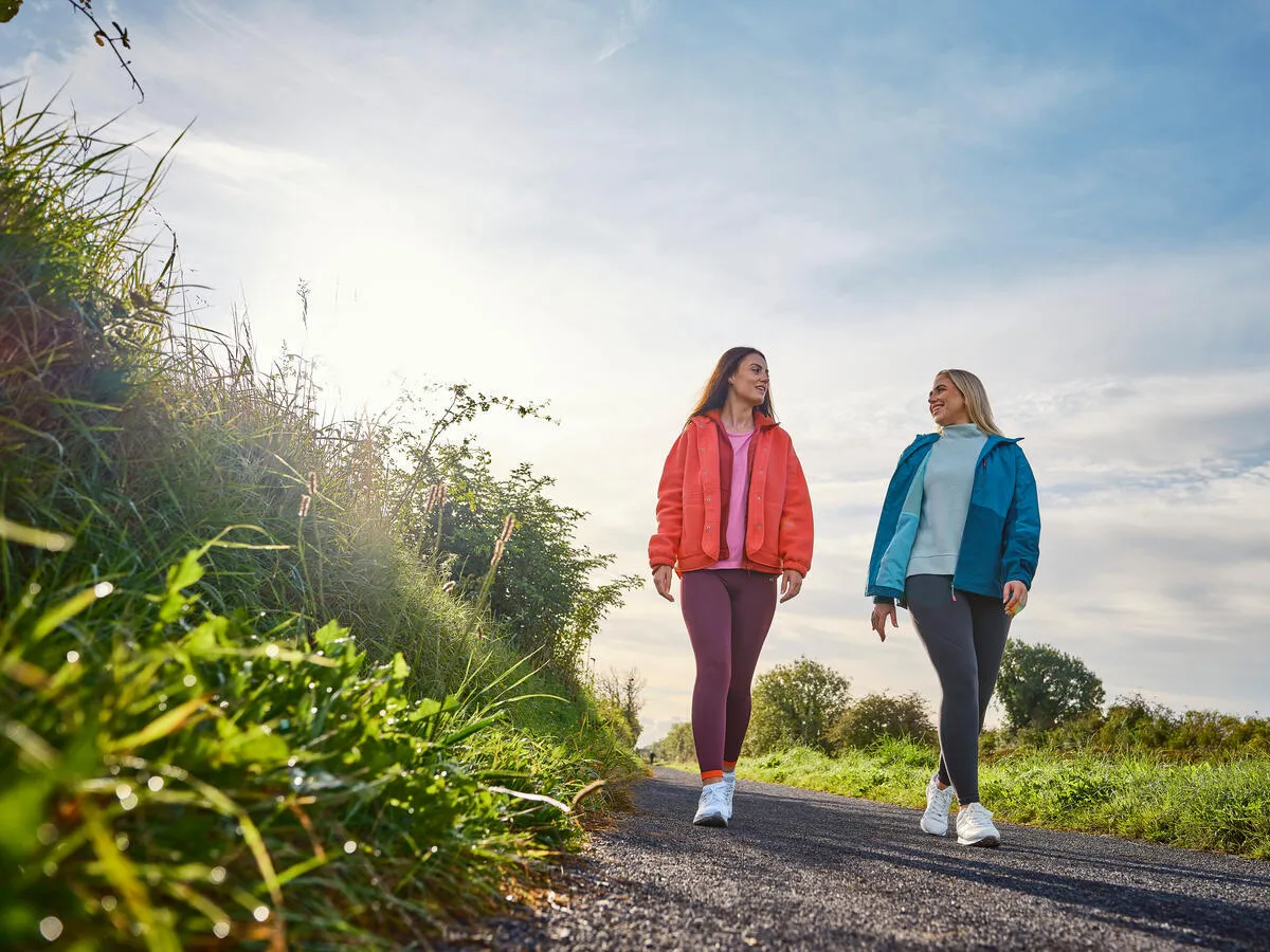 Two smiling women in jackets walking on a paved path with green grass and a blue sky.