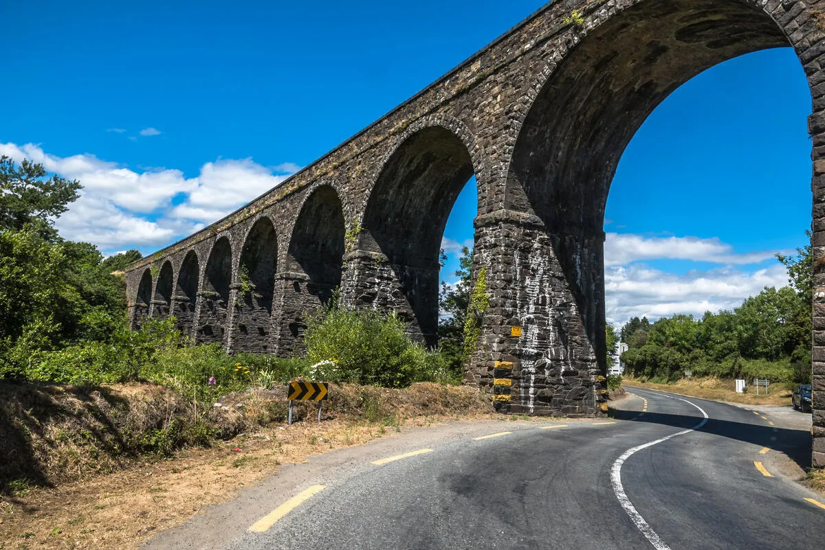 A long stone viaduct with arches spans a paved road, surrounded by green trees under a blue sky.