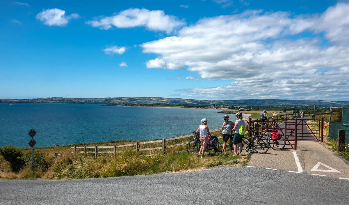 Cyclists standing near a red gate on a paved path overlooking a large body of water.